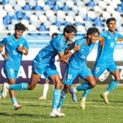 Indian football players in blue jerseys celebrate a goal during a match, with teammates running across the pitch as the crowd watches from the stands.