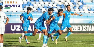 Indian football players in blue jerseys celebrate a goal during a match, with teammates running across the pitch as the crowd watches from the stands.