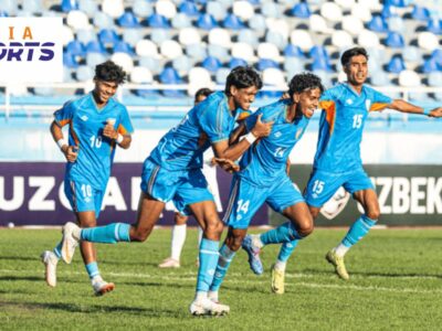 Indian football players in blue jerseys celebrate a goal during a match, with teammates running across the pitch as the crowd watches from the stands.