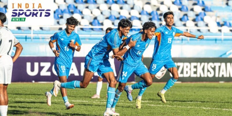Indian football players in blue jerseys celebrate a goal during a match, with teammates running across the pitch as the crowd watches from the stands.